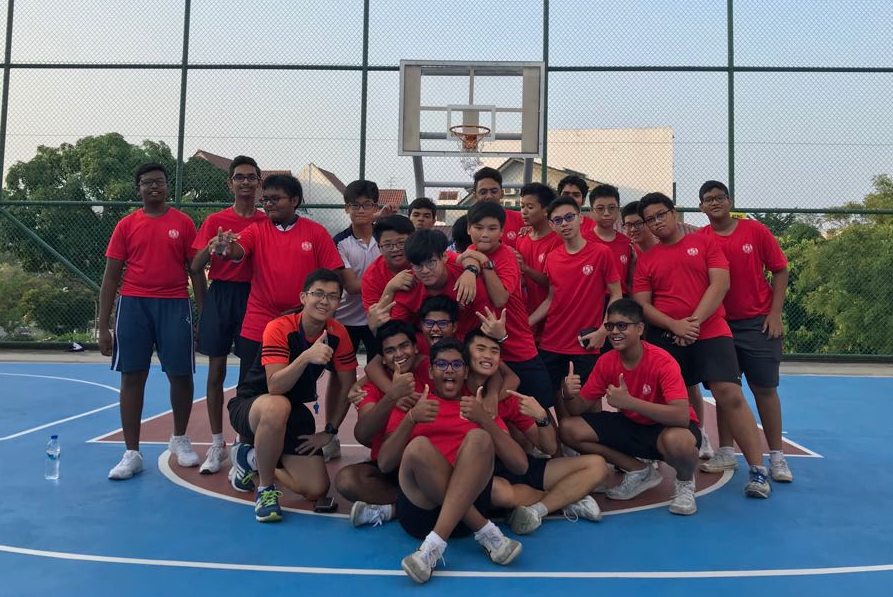 A group of teenage boys in red shirts posing together on an outdoor basketball court with a hoop visible behind them.